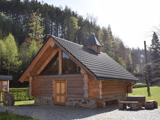 Holzkapelle mit Satteldach auf Hof, davor Bank und Holzkreuz, im Hintergrund Bäume und Wald.